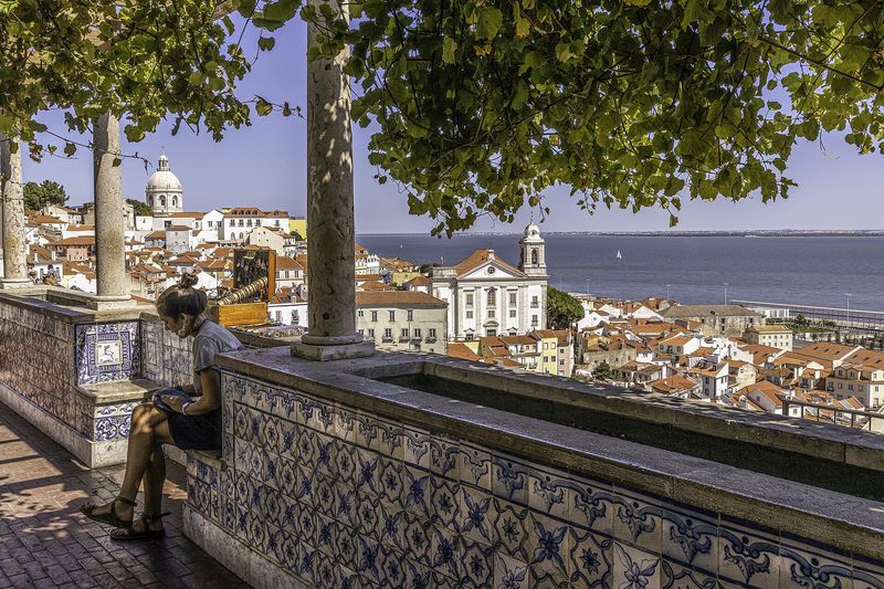 The blue-and-white azulejo-tiled terrace of the Miradouro de Santa Luzia, with its columns and shading vine, looking out over the terracotta rooftops of Alfama to the Tagus river and the dome of the National Pantheon, Lisbon