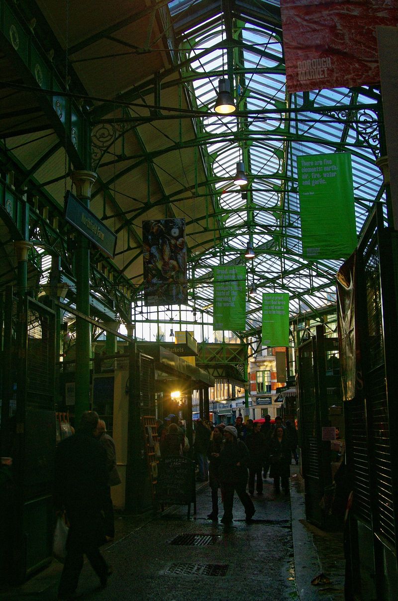 The green Victorian ironwork and glass roof of Borough Market in London, with hanging banners and shoppers below