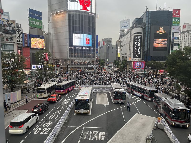 The Shibuya scramble crossing in Tokyo seen from above, crowds flooding the intersection ringed by neon screens and buses
