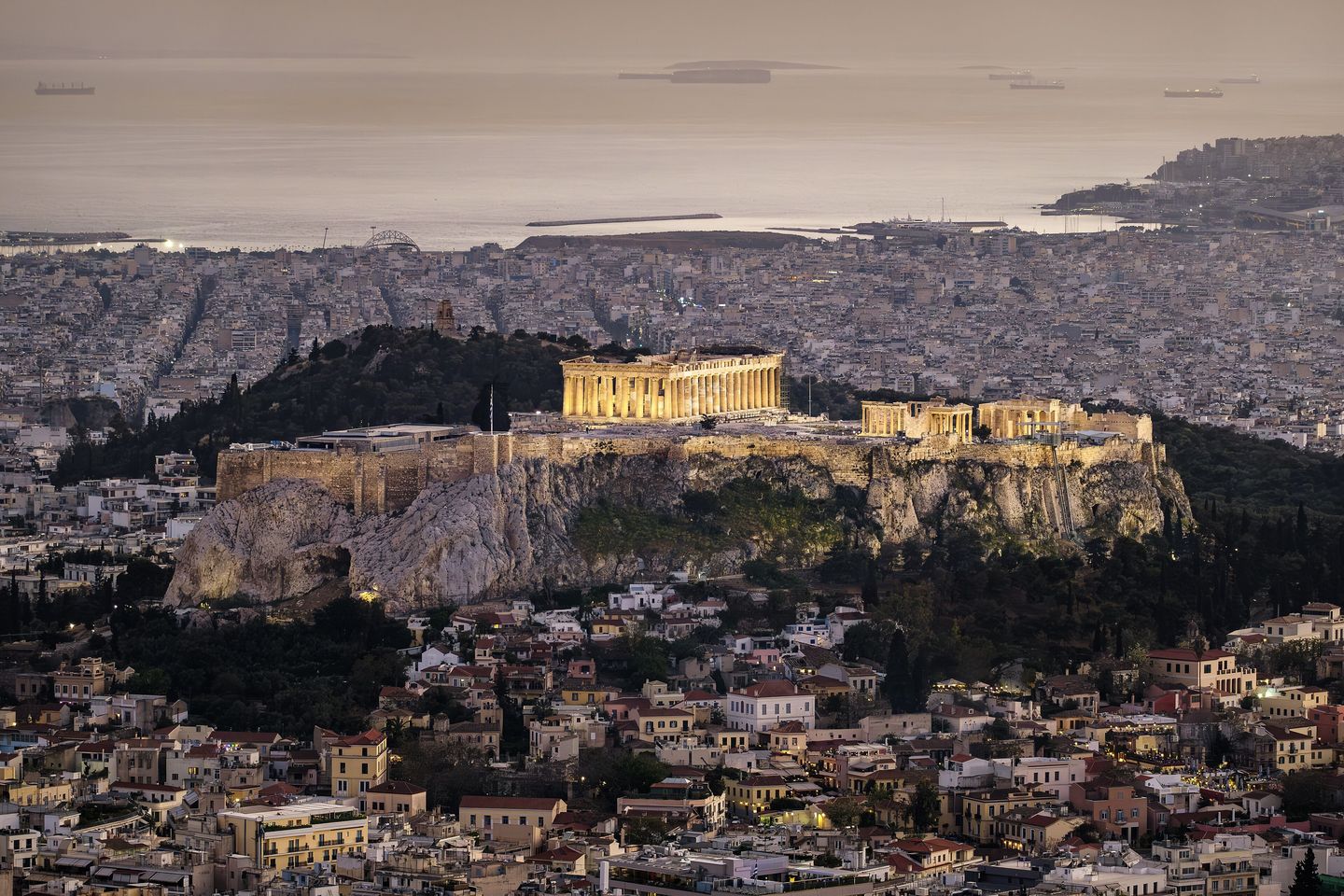 The floodlit Acropolis and Parthenon rising above the rooftops of central Athens at dusk, seen from Mount Lycabettus with the Plaka district and the Hill of the Muses visible below