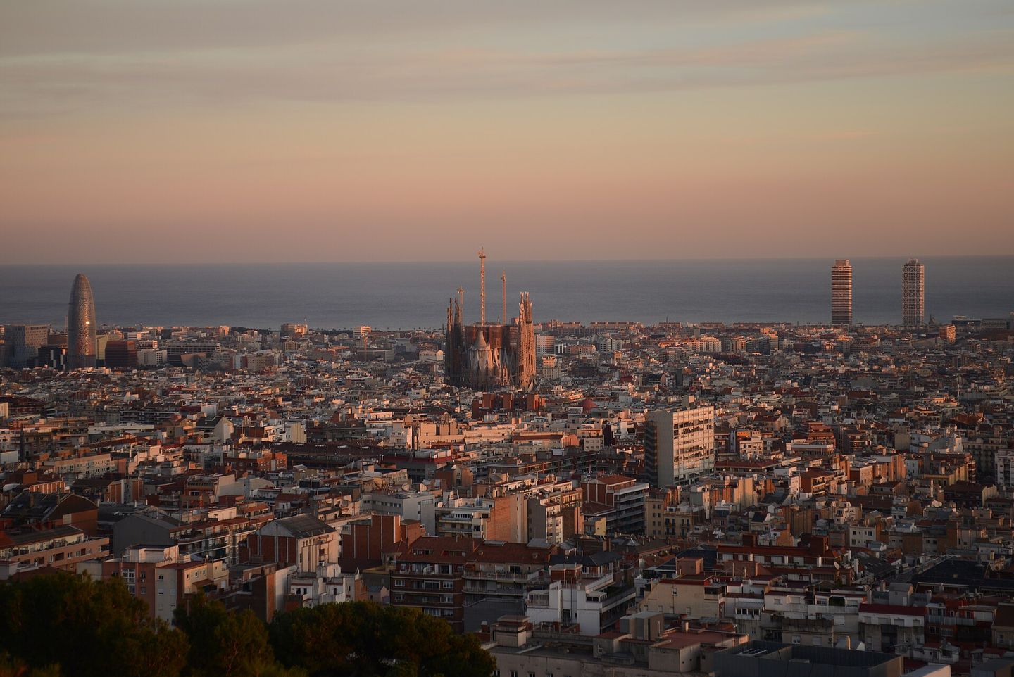 Barcelona at sunset seen from Park Güell, with the Sagrada Família rising above the Eixample rooftops, Torre Glòries at left and the Port Olímpic twin towers meeting the Mediterranean beyond
