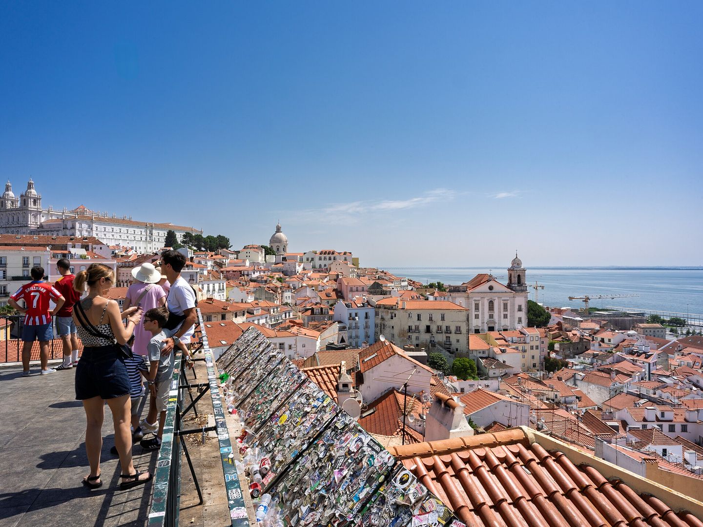 The terracotta rooftops of Alfama cascading down to the Tagus river, seen from a Lisbon miradouro, with the white twin towers of the Monastery of São Vicente de Fora and the dome of the National Pantheon rising above the old town