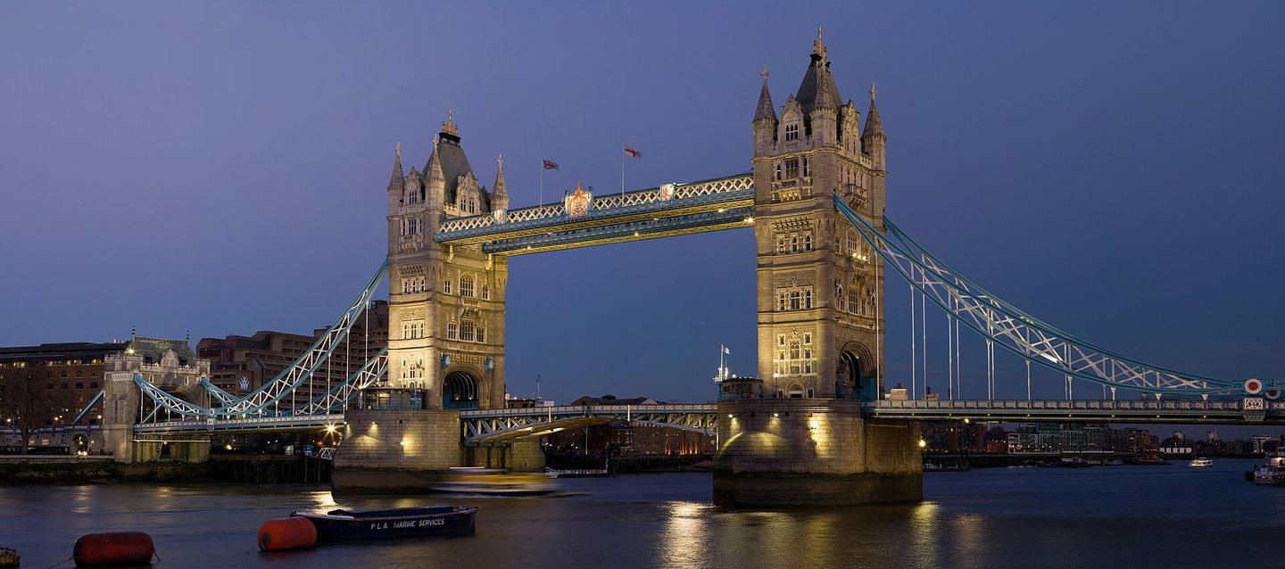 Tower Bridge illuminated at dusk over the River Thames, with its twin Gothic towers and blue suspension chains reflected in the water