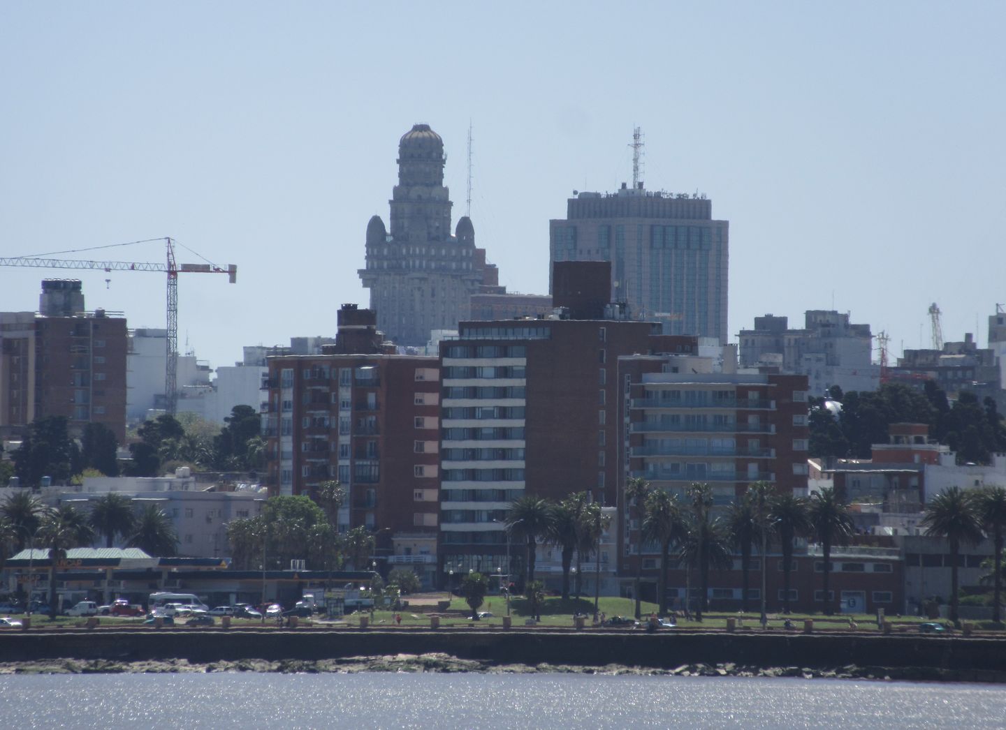 The Montevideo skyline of mixed low-rise and high-rise buildings seen across the Río de la Plata from the Rambla, with the water and a stretch of sandy shoreline in the foreground