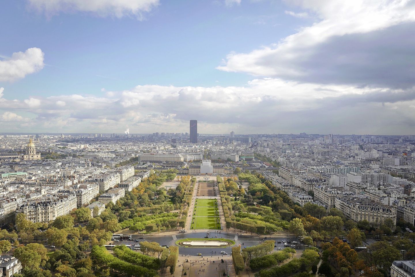 Aerial view from the Eiffel Tower down the green lawns of the Champ de Mars, over Haussmann rooftops toward the Tour Montparnasse, with the golden dome of Les Invalides at left