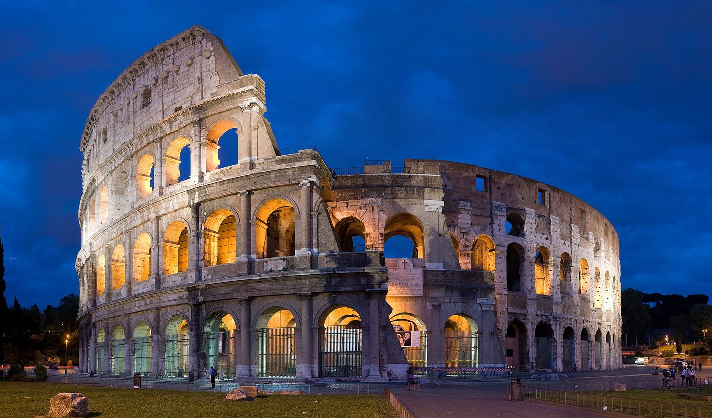 The Colosseum in Rome at dusk, its tiered arches glowing gold against a deep blue twilight sky