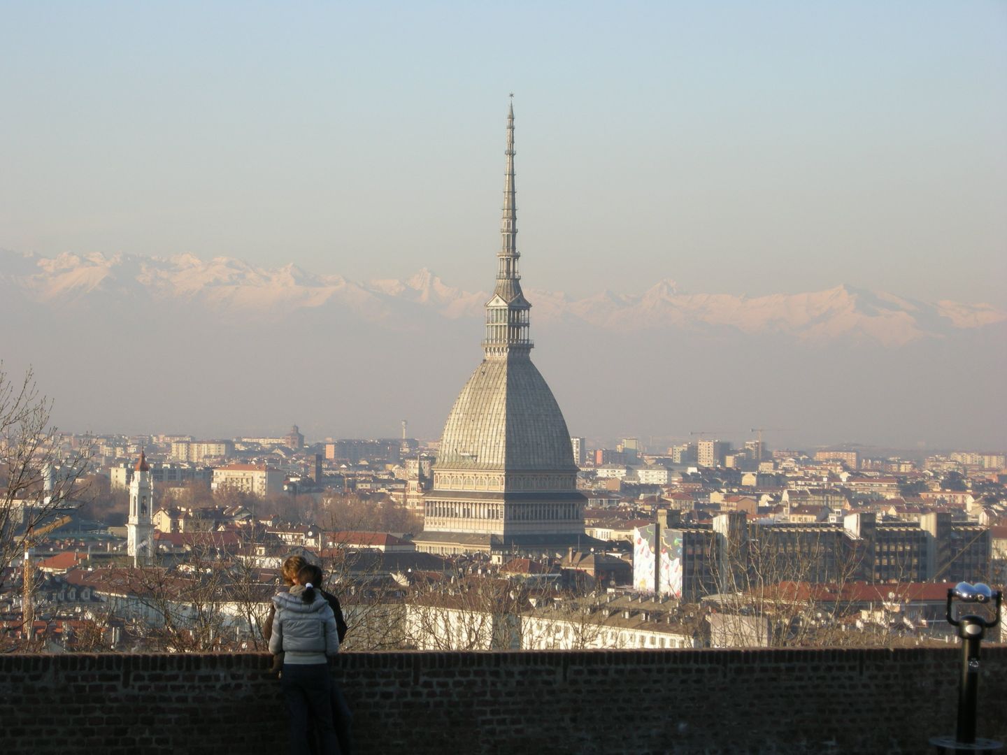 Panoramic view of Turin's rooftops from the Cappuccini hill, with the tall spire of the Mole Antonelliana rising above the Baroque cityscape and the Alps faint on the horizon