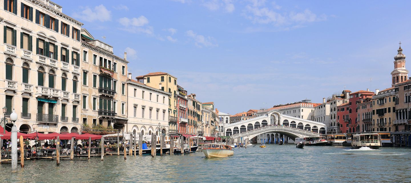 Panoramic view of the Grand Canal and the Rialto Bridge in Venice, with gondolas and vaporetti on the water and pastel-coloured palazzo facades lining both banks